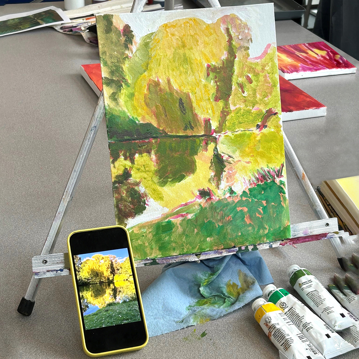 Painting of a colorful landscape painting on a tabletop easel referencing a landscape photo on a smartphone screen