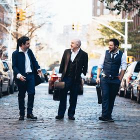 The Secret Trio - three musicians carrying instruments in cases down a city street